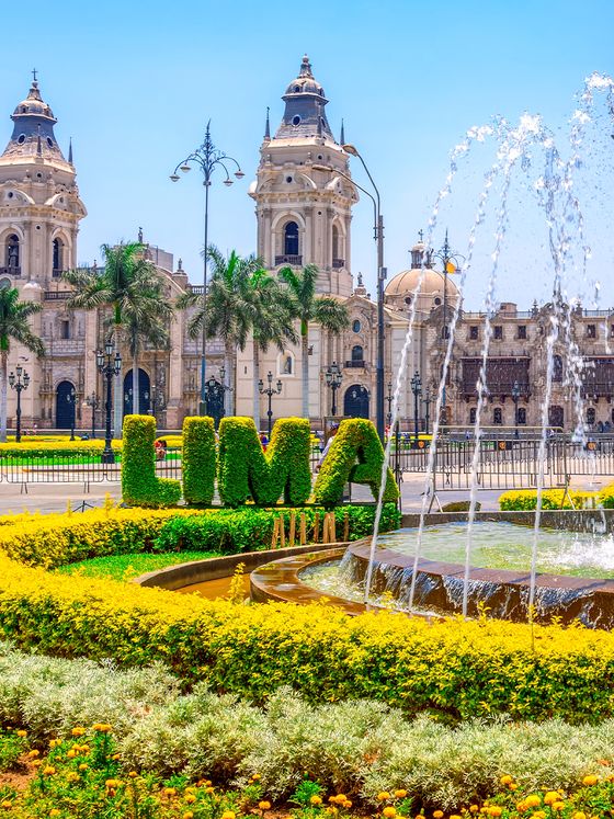 View of Lima's main square with colonial architecture, lush greenery, "LIMA" sign, and a fountain in the foreground under a clear blue sky.