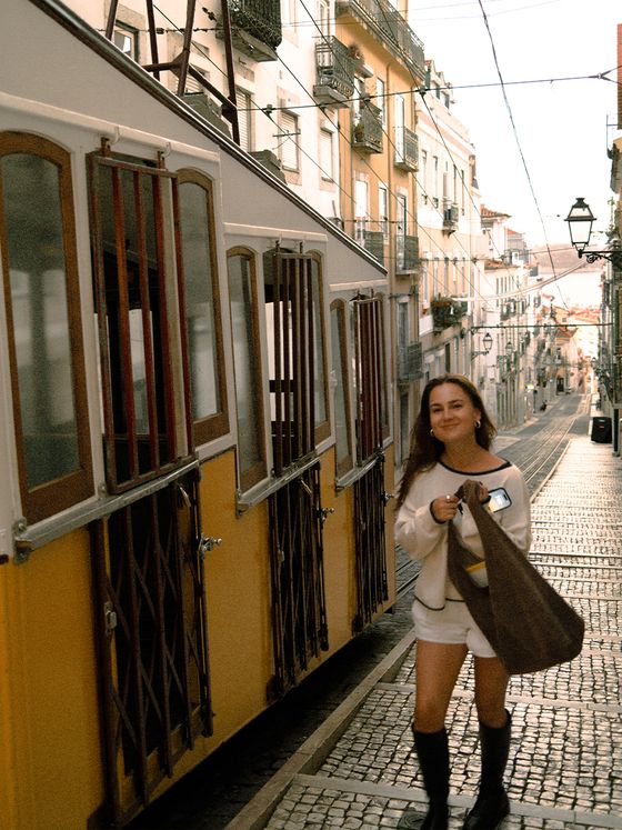 A woman in a white outfit and black boots walks on a cobblestone street beside a yellow tram in a European city.