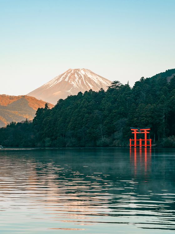Snow-capped Mount Fuji behind a forest and a red torii gate reflecting on a calm lake under a clear blue sky.