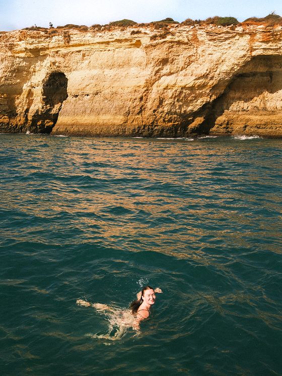 Person swimming in clear blue water near large, rugged cliffs under a bright sky.