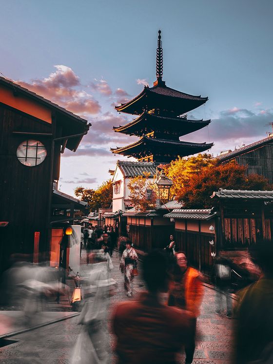 A bustling street scene with blurred motion, featuring a traditional Japanese pagoda under a sunset sky, surrounded by wooden buildings and autumn trees.