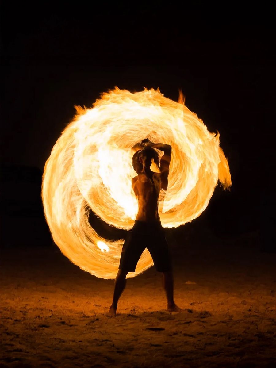 A person skillfully performs fire dancing on a dark beach, creating a vibrant swirling pattern of flames around their body.