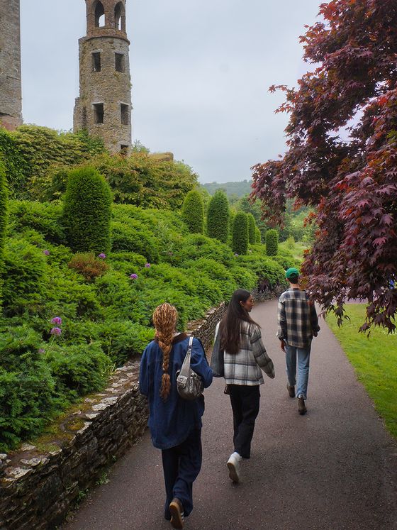 Three people walking on a path beside lush greenery and an old stone tower.