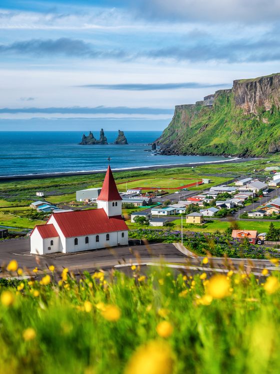 Scenic view of a small coastal village with a red-roofed church, dramatic cliffs, and ocean in the background, surrounded by yellow flowers.