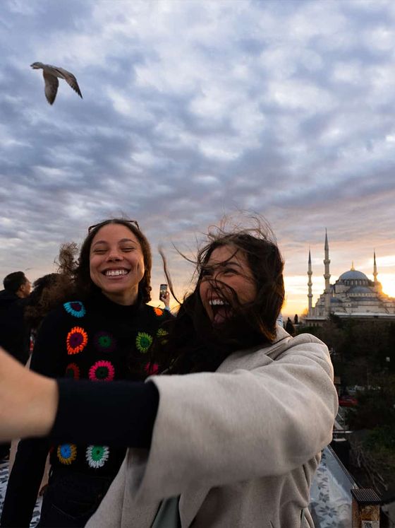 Two women smiling and taking a selfie at sunset with the Blue Mosque in the background and a bird flying nearby under a cloudy sky.
