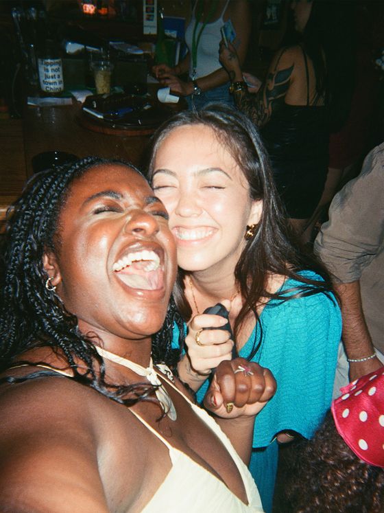 Two people laughing joyfully in a lively social setting, surrounded by others. One is wearing a turquoise top, both showing joyous expressions.