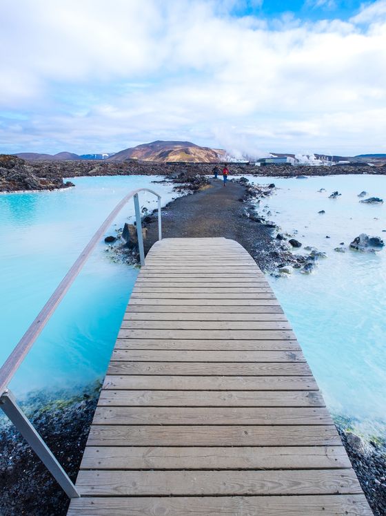 Wooden bridge over serene blue geothermal waters with distant figures and mountainous landscape under a cloudy sky.