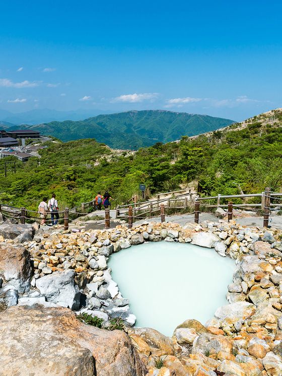 A small, heart-shaped hot spring surrounded by rocks, set in a mountainous landscape under a clear blue sky, with people walking nearby.