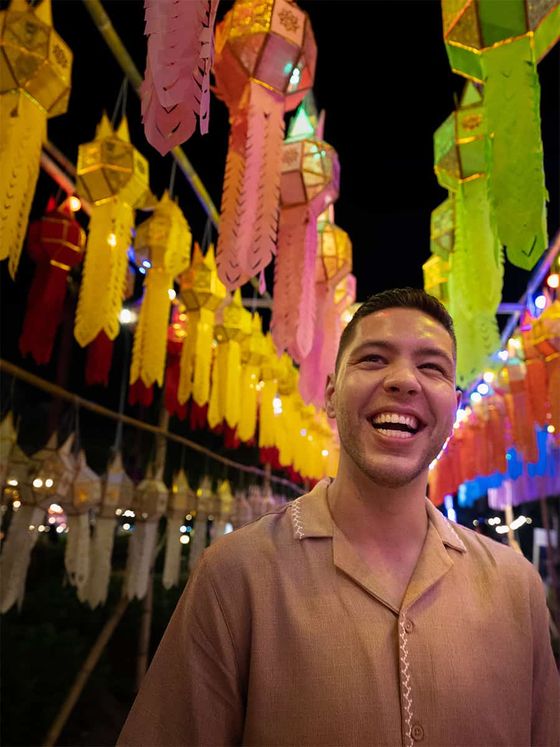 Smiling man stands under vibrant, colorful lanterns at night, wearing a beige shirt.