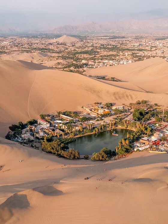 Aerial view of the Huacachina oasis surrounded by sand dunes, with greenery and buildings, set against a distant mountainous landscape.