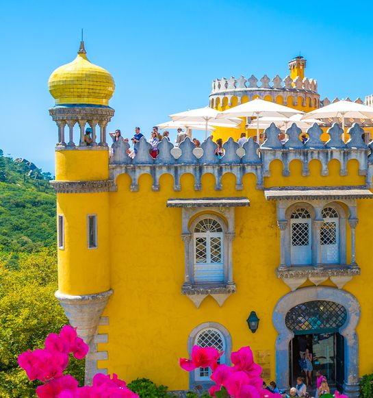 Colorful yellow castle with domed turret, surrounded by greenery and vibrant pink flowers, under a clear blue sky.