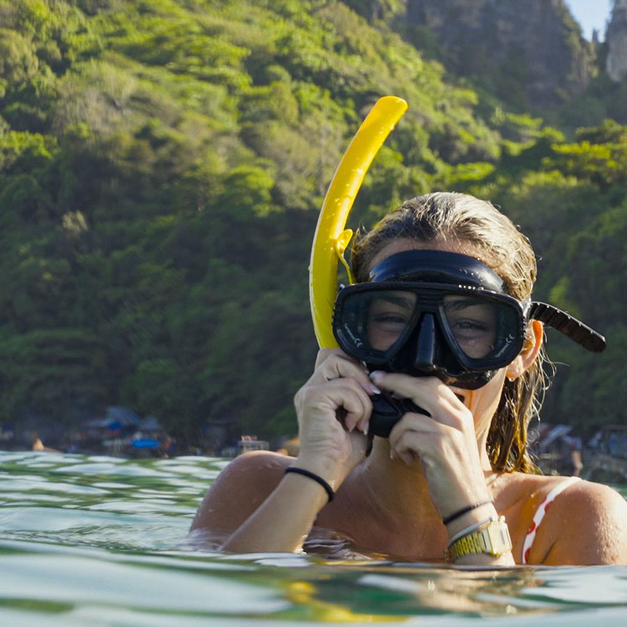 Swimmer in Thailand with black snorkel mask and yellow snorkel emerging from water, lush forested cliffs in the background.