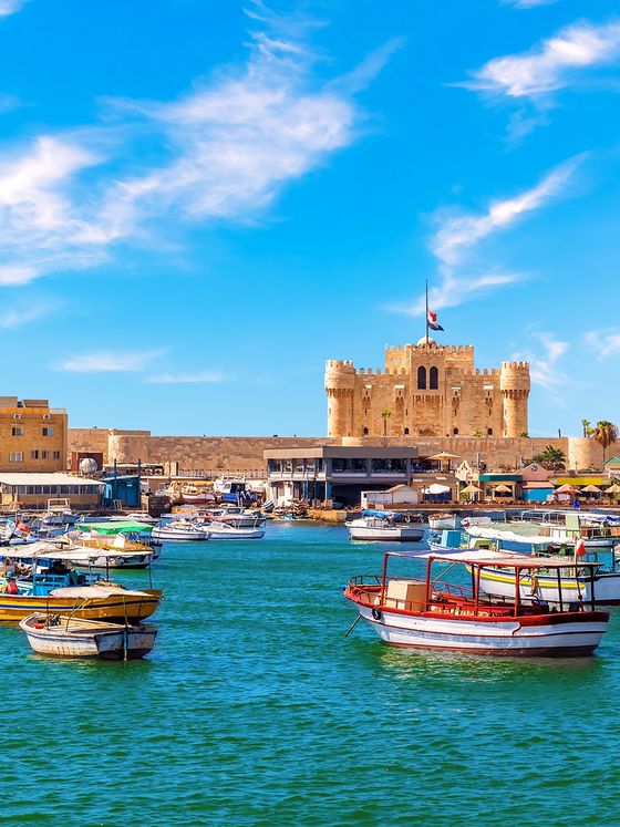 Boats anchored in a vibrant blue harbor with a historic fortress in the background under a bright, partly cloudy sky.