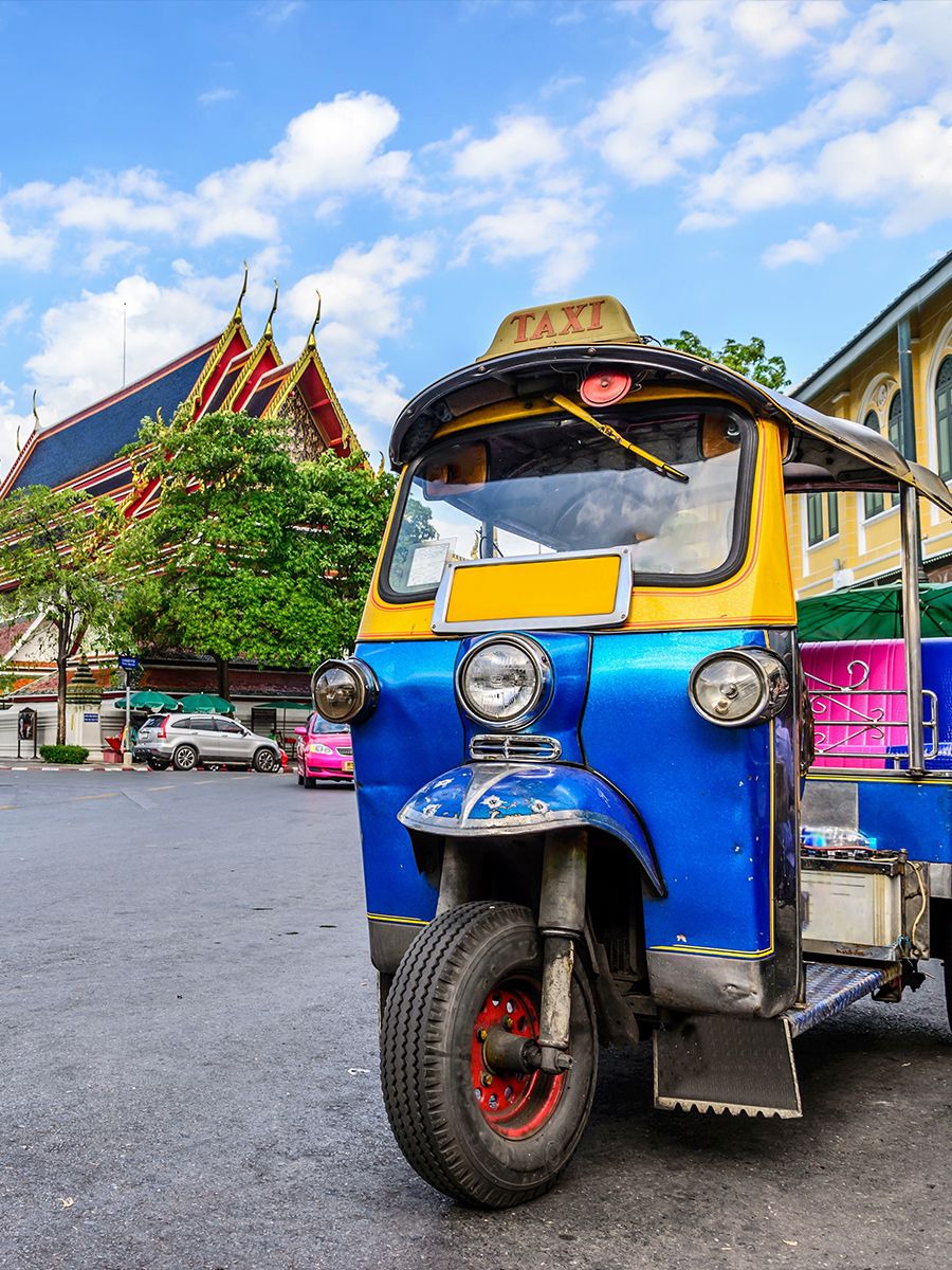 A vibrant blue and yellow tuk-tuk taxi is parked on a street with traditional Thai architecture and a clear blue sky in the background.
