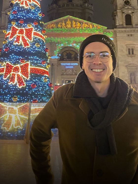 Man smiling in front of a brightly lit Christmas tree and a building with festive decorations at night.