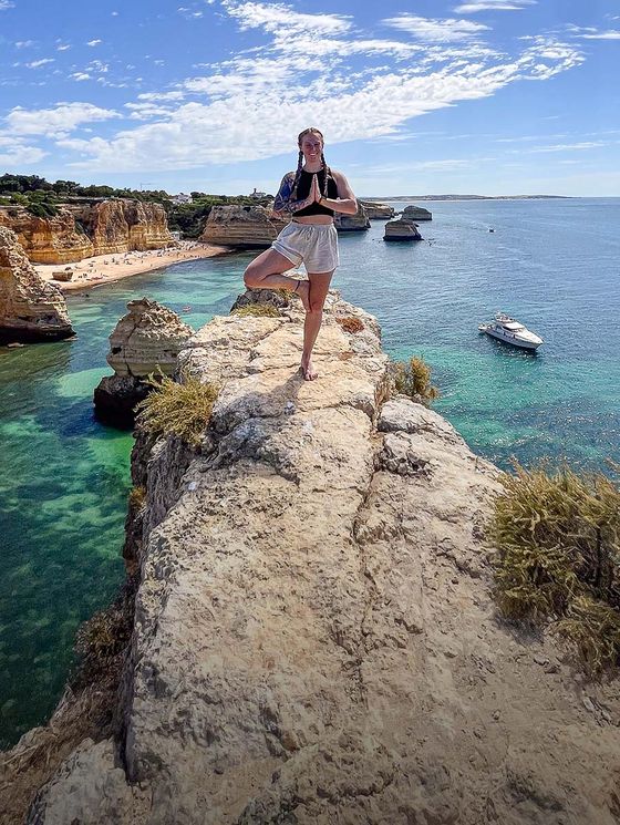 Person in a yoga pose on a scenic cliff overlooking turquoise waters, with boats and rock formations in the background under a blue sky.