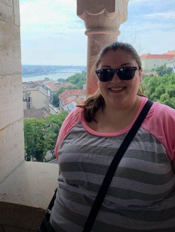 Smiling person wearing sunglasses and a striped shirt stands by a stone column with a city and river view in the background.