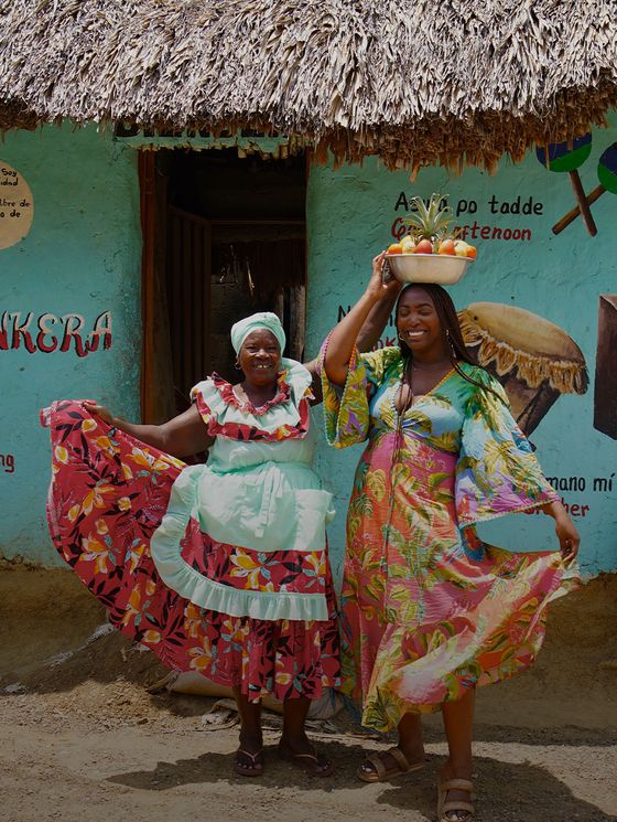Two women in colorful dresses smile outside a building with a thatched roof. One holds her skirt, and the other balances a basket on her head.