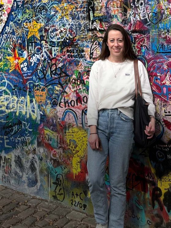 Smiling woman in casual clothes stands in front of a colorful, graffiti-covered wall.