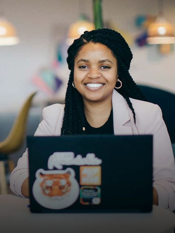 Smiling woman with braided hair working on a laptop at a desk in a modern, colorful office setting.