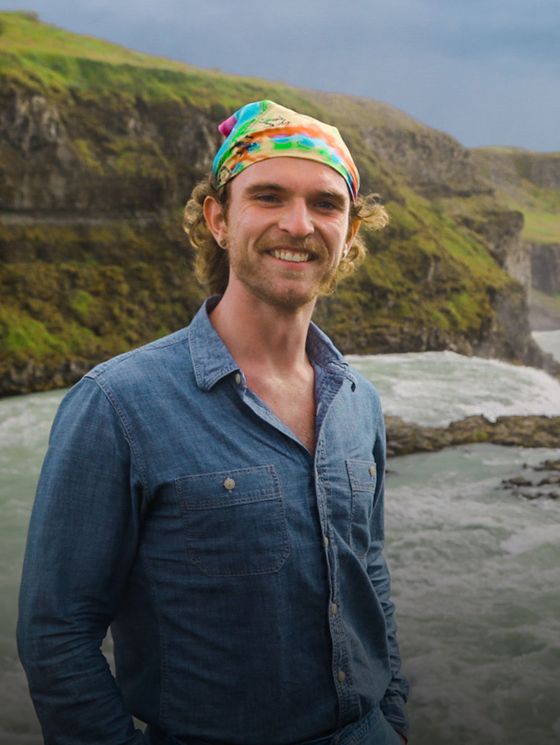 Smiling man in a denim shirt and colorful bandana stands by a river with rocky cliffs and green landscape in the background.