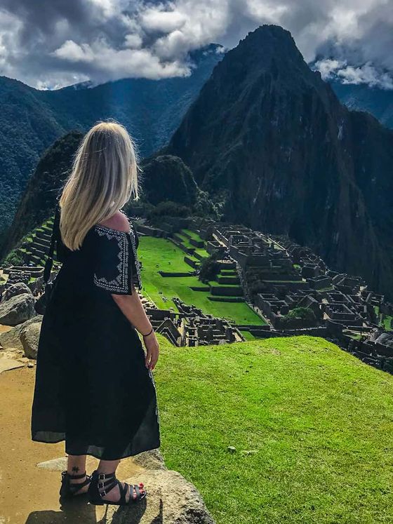 A woman with long blonde hair and a black dress stands on a hill overlooking the ancient ruins of Machu Picchu under a cloudy sky.