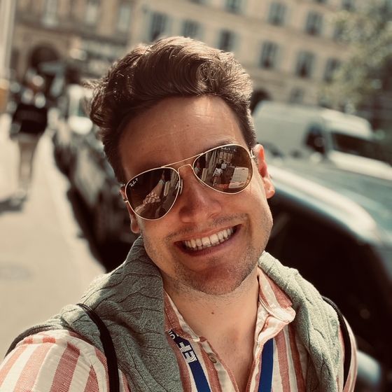 Person smiling in a striped shirt and sunglasses, standing on a sunny street with cars and a building in the background.