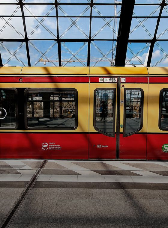 A yellow and red train stopped at a glass-roofed station; a person is reflected in the train's windows, and the sky is partly cloudy.