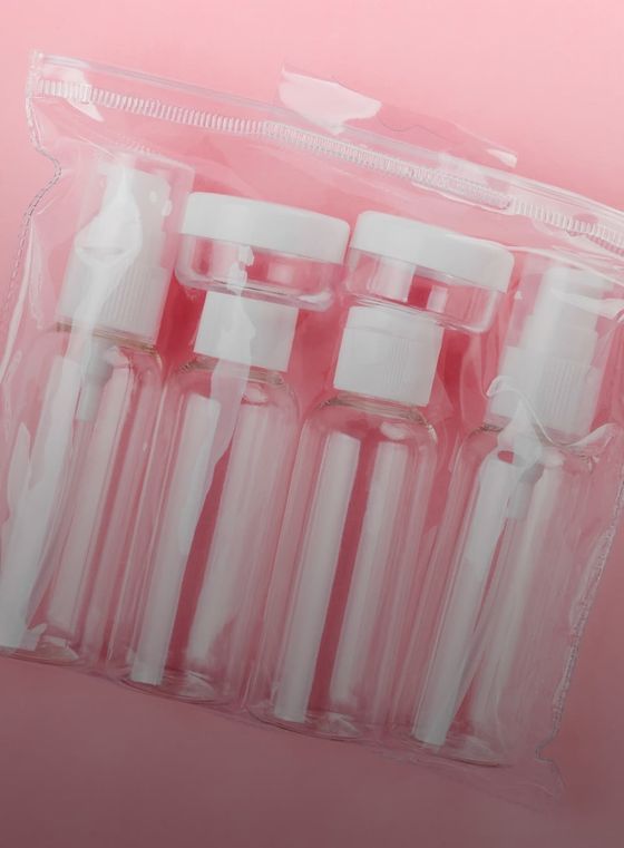 Transparent toiletry bottles with white caps in a clear plastic zippered pouch, set against a pink background.