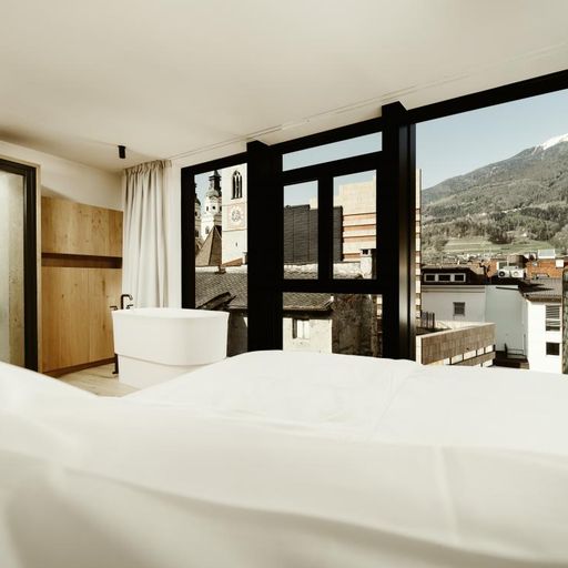 Modern hotel room with a large bed, freestanding bathtub, and floor-to-ceiling windows offering a scenic mountain and town view.