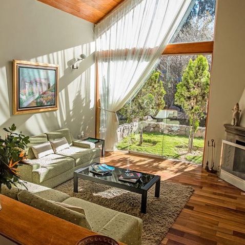 Sunlit living room with large windows, green sofas, a glass coffee table, and a fireplace. Outside, a garden with trees is visible.