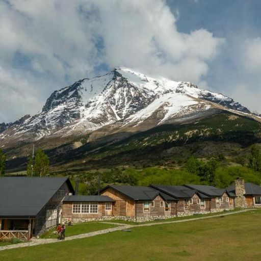 Rustic wooden cabins sit at the base of a snow-capped mountain under a partly cloudy sky, with lush greenery in the foreground.