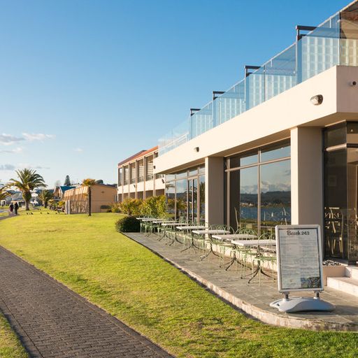 Modern lakeside building with glass balconies, outdoor seating, and a paved path on a sunny day. Palm trees and water are in the background.