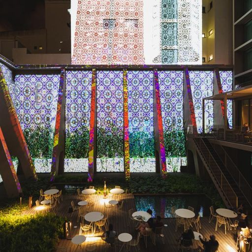 Outdoor dining area with tables and chairs, surrounded by illuminated, patterned walls at night, creating a vibrant and lively atmosphere.