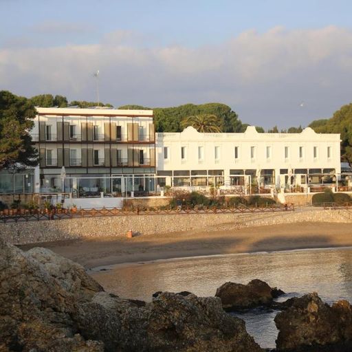 A white beachfront hotel with balconies overlooks a sandy beach and calm sea, surrounded by trees under a partly cloudy sky.