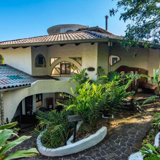 A two-story house with a tiled roof, surrounded by lush tropical plants and a cobblestone path under a clear blue sky.