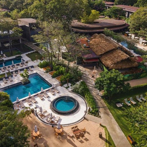 Aerial view of a resort with multiple pools, lounge chairs, a circular hot tub, thatched-roof building, and lush greenery.