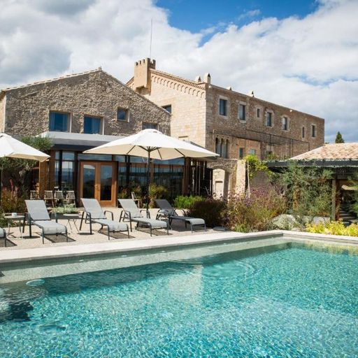 Stone villa with a large swimming pool, sun loungers, and umbrellas under a partly cloudy sky, surrounded by greenery.