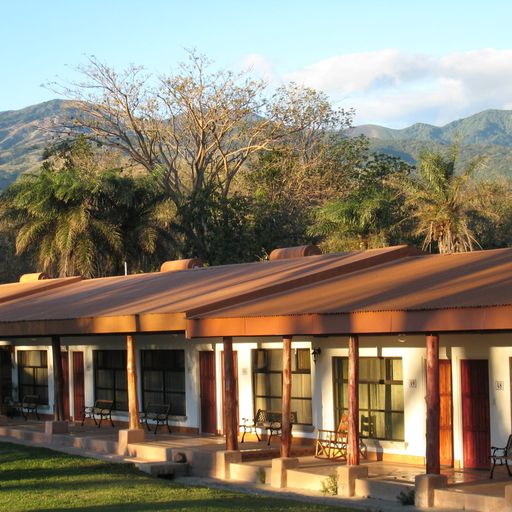 A row of rustic cabins with wooden columns, set against a backdrop of lush trees and distant mountains under a clear blue sky.