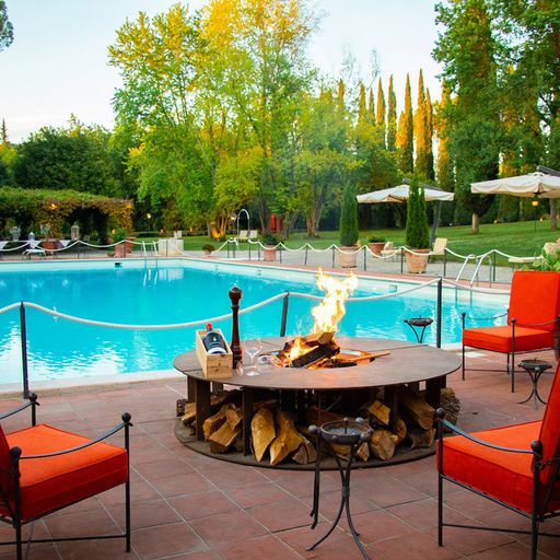 Outdoor pool area with a central fire pit surrounded by red chairs. Lush greenery and umbrellas in the background create a serene atmosphere.