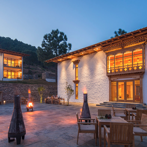 A serene courtyard at dusk with two modern buildings featuring warm lighting, wooden accents, and outdoor seating around lit fire pits.