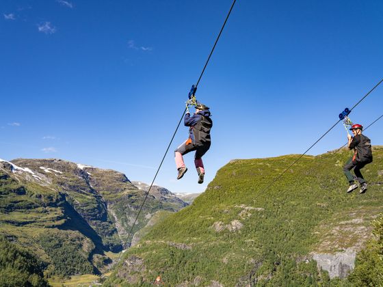 Two people zip-lining across a lush valley with mountainous terrain and a clear blue sky in the background.