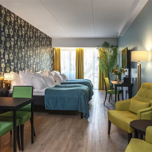 Hotel room with two beds, green chairs, a desk, and a large mirror. Trophy-patterned wallpaper and a window with yellow curtains brighten the space.