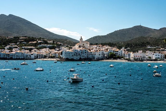 Coastal town with white buildings by a blue sea, surrounded by mountains. Boats float in the water under a clear sky.