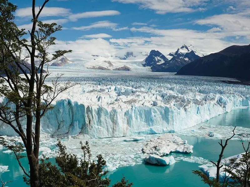 A stunning glacier with icy blue colors extends into a turquoise lake, surrounded by jagged mountains and framed by sparse trees under a partially cloudy sky.