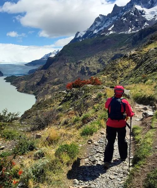 A hiker in red gear walks along a scenic mountain trail overlooking a turquoise lake and snow-capped peaks under a partly cloudy sky.