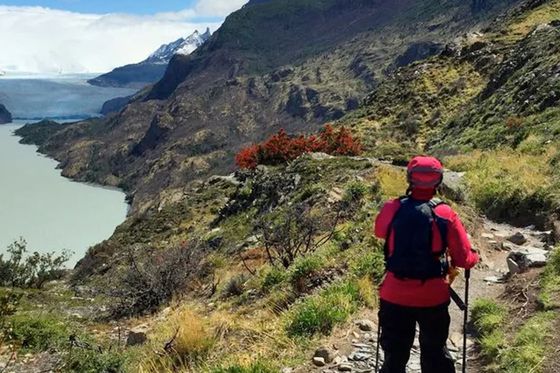 A hiker in red gear walks along a scenic mountain trail overlooking a turquoise lake and snow-capped peaks under a partly cloudy sky.
