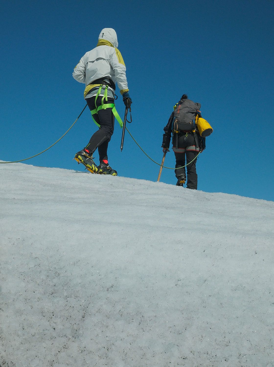 Two climbers roped together, wearing climbing gear and backpacks, traverse a snow-covered slope under a clear blue sky.