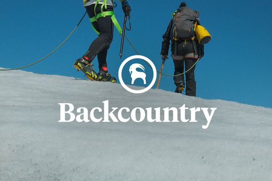 Two climbers with backpacks and ice picks ascend a snowy slope under a clear blue sky, connected by a safety rope. "Backcountry" logo is visible.