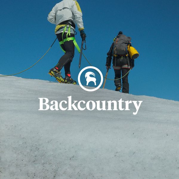 Two climbers with backpacks and ice picks ascend a snowy slope under a clear blue sky, connected by a safety rope. "Backcountry" logo is visible.